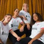 Four female football players in white Liverpool jerseys pose together against a gold curtain backdrop, looking confident and united.