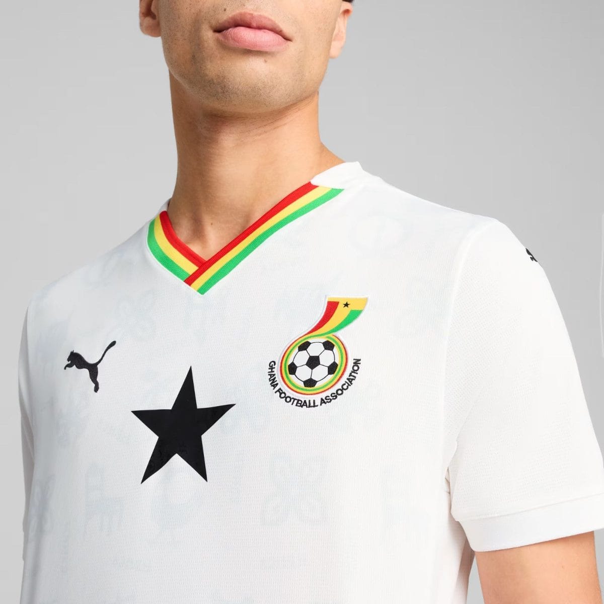 Close-up of a white football jersey featuring a rainbow-trim V-neck, the Ghana Football Association crest, and a black star on the chest under a gray background.