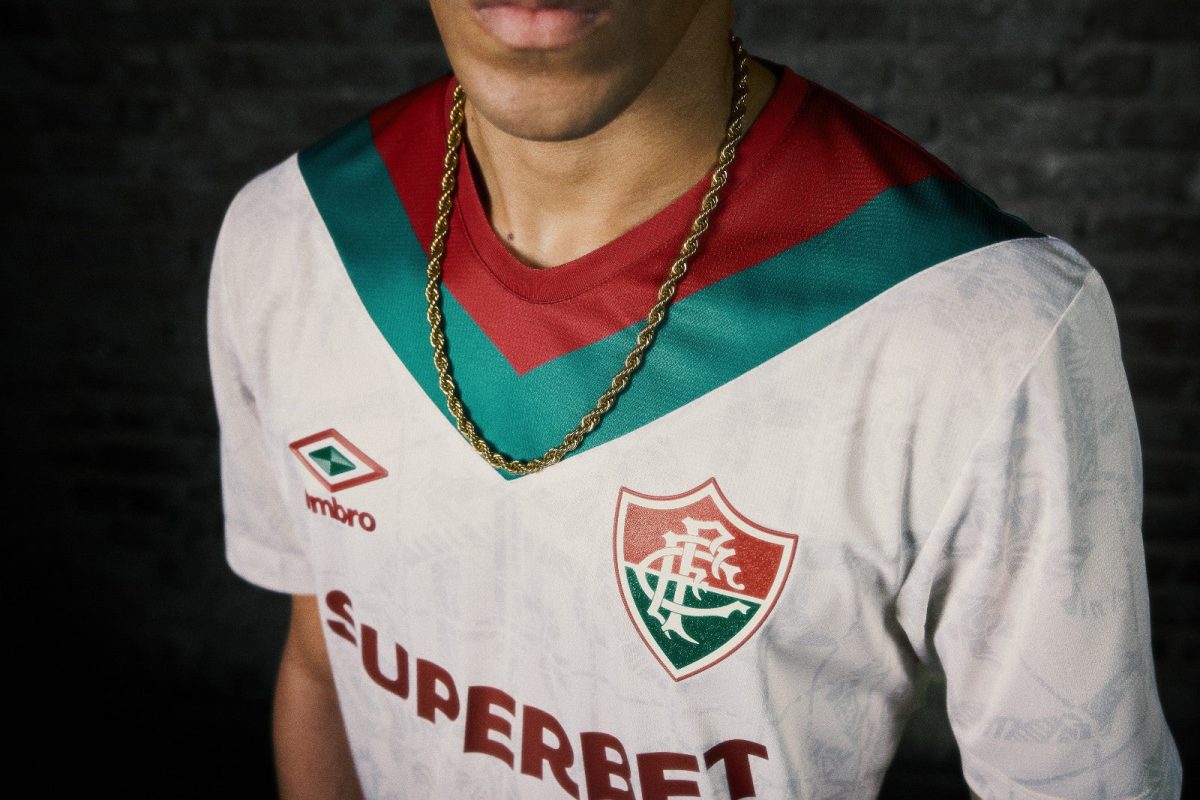 Close-up of a person wearing a white soccer jersey with red and green V-neck, a gold chain, and crest on a dark background.