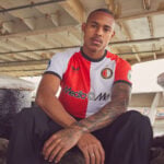 Portrait of a man with a tattooed arm, seated under a bridge, wearing a red-and-white F.C. Feyenoord jersey with a 'Media' sponsor logo.