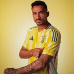 Man with arms crossed wearing a yellow Juventus football jersey, tattooed forearms, against a yellow background.