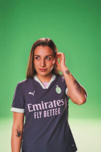 Portrait of a woman in a dark purple soccer jersey with the Emirates Fly Better logo, against a green screen backdrop, adjusting her hair.