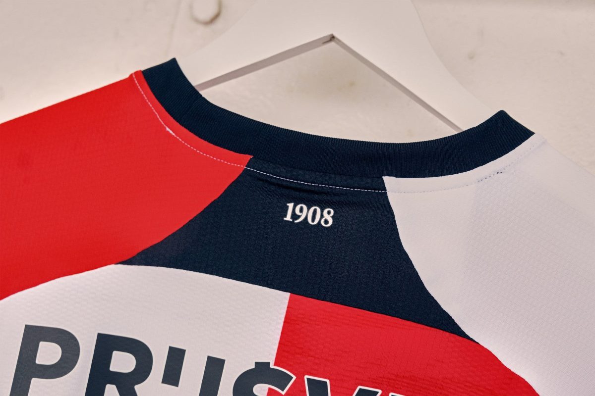 Close-up of a color-blocked jersey (red, navy, white) hanging on a hanger, with the year '1908' printed near the neckline.