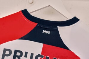 Close-up of a color-blocked jersey (red, navy, white) hanging on a hanger, with the year '1908' printed near the neckline.