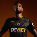 Portrait of a man wearing a black and gold football jersey with a wolf crest, standing against an orange background.