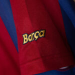 Close-up of a red FC Barcelona jersey sleeve with a yellow embroidered 'Barca' logo on blue fabric accents.