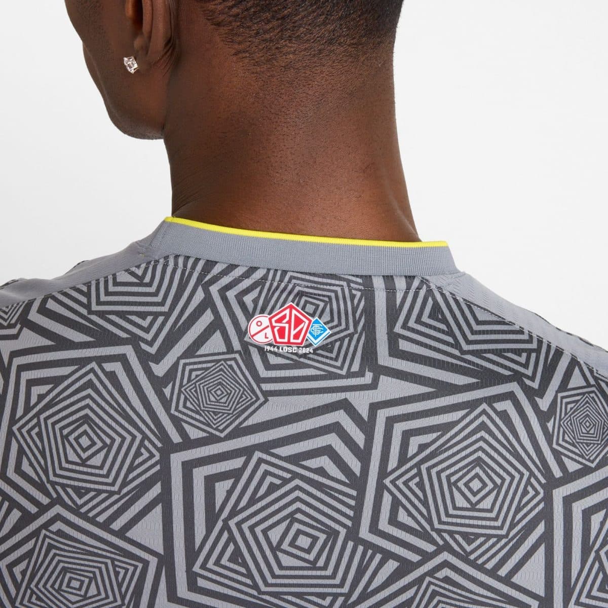 Back of a person wearing a gray geometric-patterned shirt; a small red-blue emblem sits at the collar area, with an earring visible on the ear.