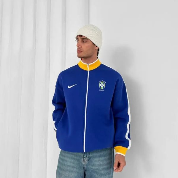 Man wearing a royal blue Brazil jacket with a yellow collar and white zipper, paired with a cream knit beanie and jeans, posing against a white backdrop.