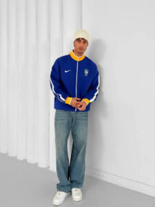Man wearing a blue Brazil jacket with yellow cuffs, light beanie, jeans, and white sneakers, standing against a white wall.