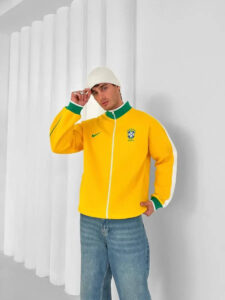 Man wearing a bright yellow Brazil track jacket with green collar, tipping a white knit beanie while leaning against a white wall.
