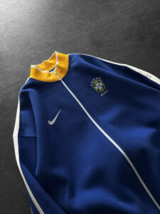 Blue Brazil national team track jacket with yellow collar, white zipper, and Nike swoosh on a dark background.