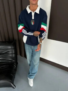 Person standing indoors in a navy polo with an Italian crest, color-block sleeves, jeans and white sneakers, arms crossed.