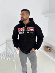 Man standing in a white room wearing a black zip-up hoodie with racing sponsor patches on the chest, looking to the left.