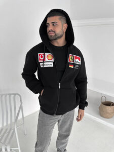 Man wearing a black zip-up hoodie with sponsor patches on the chest, looking to his left in a white room.