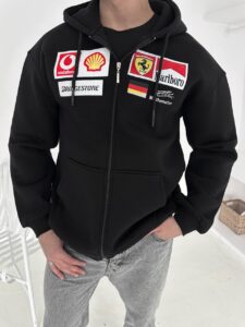 Person wearing a black zip-up hoodie with racing sponsor patches (Vodafone, Shell, Ferrari, Marlboro, Bridgestone) on the chest and a German flag patch, standing with hands in pockets.