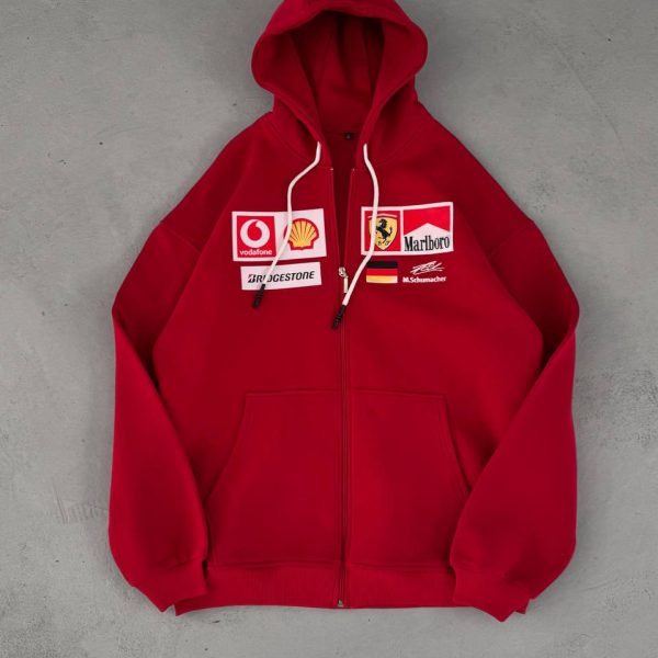Red zip-up hoodie with white drawstrings lying on a gray concrete surface, featuring sponsor patches on the chest area (Vodafone, Shell, Marlboro, etc.).