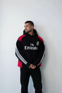 photo_2025-11-09_15-10-55 | UAE SPORT STORE Man in a black Real Madrid hoodie with red panels, looking to the left, standing against a white wall.