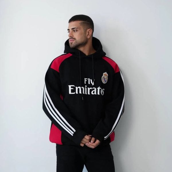 Man in a black Real Madrid hoodie with red panels, looking to the left, standing against a white wall.