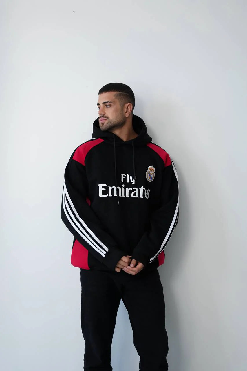 photo_2025-11-09_15-10-55 | UAE SPORT STORE Man in a black Real Madrid hoodie with red panels, looking to the left, standing against a white wall.