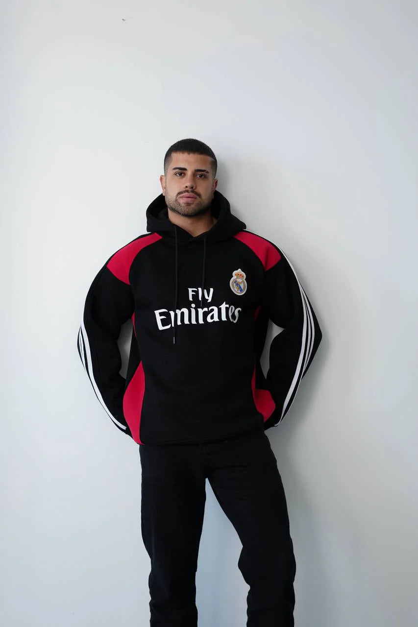 photo_2025-11-09_15-10-56_2 | UAE SPORT STORE Man posing in a black Real Madrid hoodie with red panels and 'Fly Emirates' logo, against a white wall