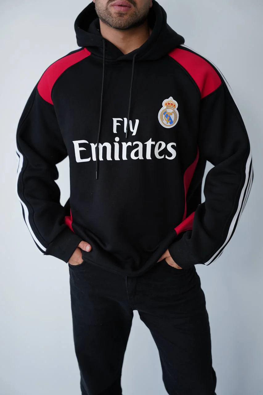 photo_2025-11-09_15-10-58 | UAE SPORT STORE Man wearing a black Real Madrid hoodie with red accents and the 'Fly Emirates' logo, standing with hands in pockets.