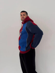 Man in profile wearing a blue and maroon color-block hoodie, hands behind his back, against a plain wall.