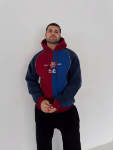 Man posing against a plain white wall in a red-and-blue color-block hoodie with a club crest on the chest, hands clasped in front.