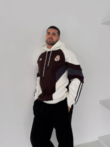 Man posing against a white wall in a color-block Real Madrid hoodie with a chest logo, hands in pockets, relaxed stance
