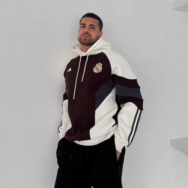 Man posing against a white wall in a color-block Real Madrid hoodie with a chest logo, hands in pockets, relaxed stance