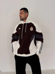 Man wearing a white and dark brown Real Madrid hoodie with the club crest on the left chest, standing against a plain white wall and looking to the side.
