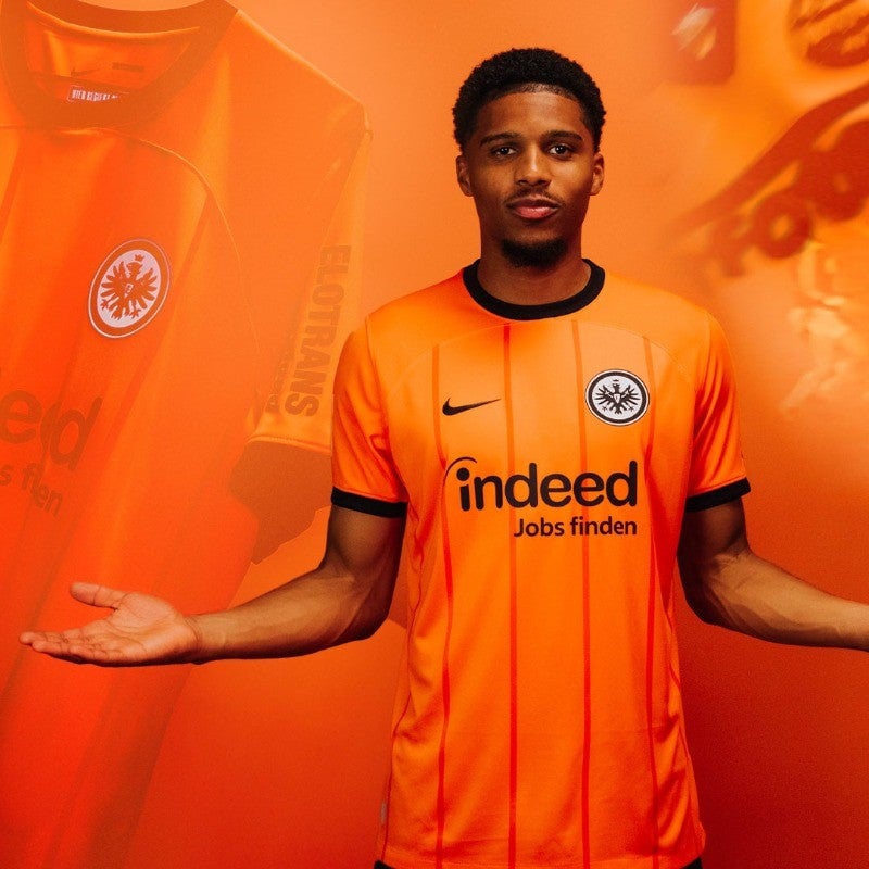pnY11tAq2rLV8QX | UAE SPORT STORE Male footballer wearing a bright orange jersey with sponsor text 'indeed Jobs finden' and team crest, standing with arms open against an orange backdrop.