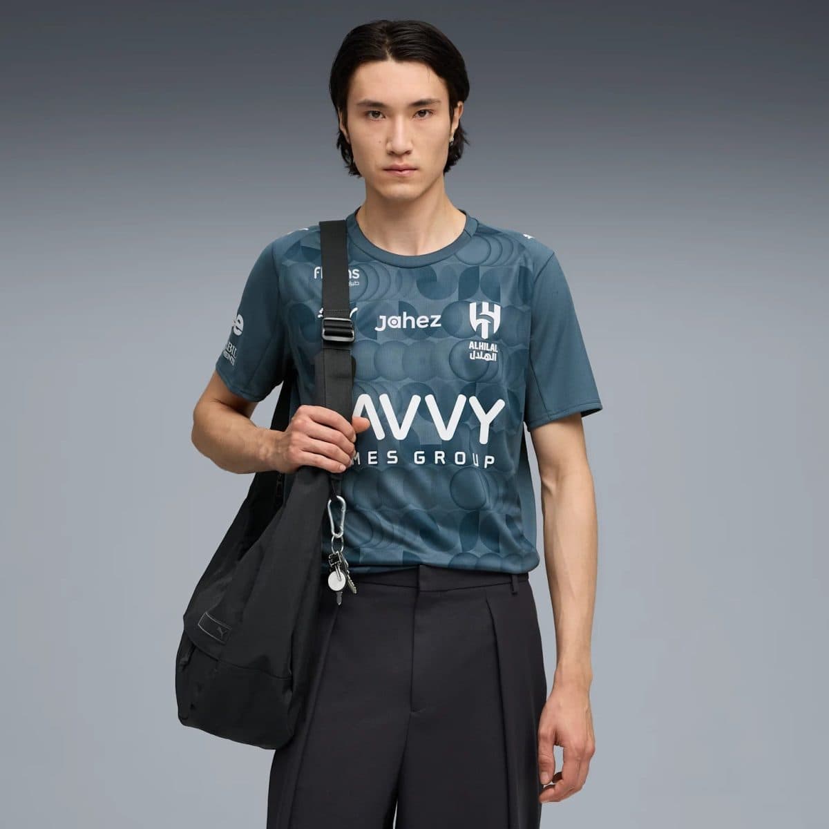 Young man in a teal athletic shirt with logos, crossbody bag over his shoulder, standing against a gray gradient background.