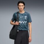 Young man in a teal athletic shirt with logos, crossbody bag over his shoulder, standing against a gray gradient background.