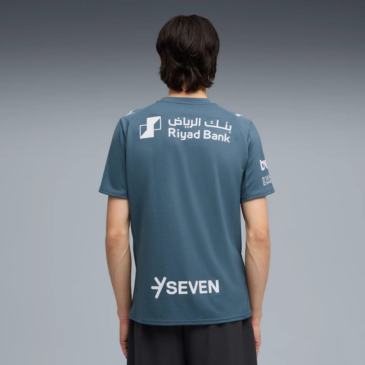 Back view of a person wearing a blue T-shirt with Riyad Bank logo and 'SEVEN' text on the lower back area.