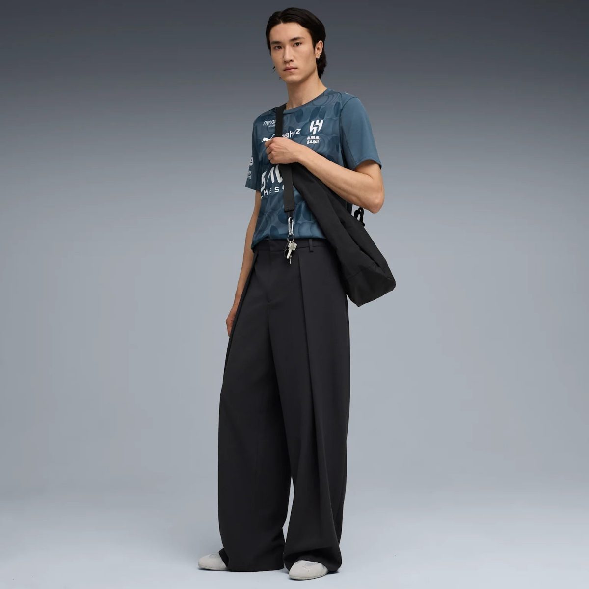 Male model wearing a teal patterned T-shirt, oversized black wide-leg pants, white sneakers, and a black shoulder bag with keys hanging from the strap.