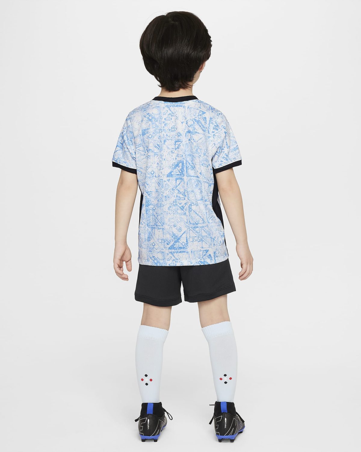 Child seen from the back wearing a light blue patterned sports jersey, black shorts, white knee-high socks with small red/black dots, and black-and-blue cleats with blue accents.