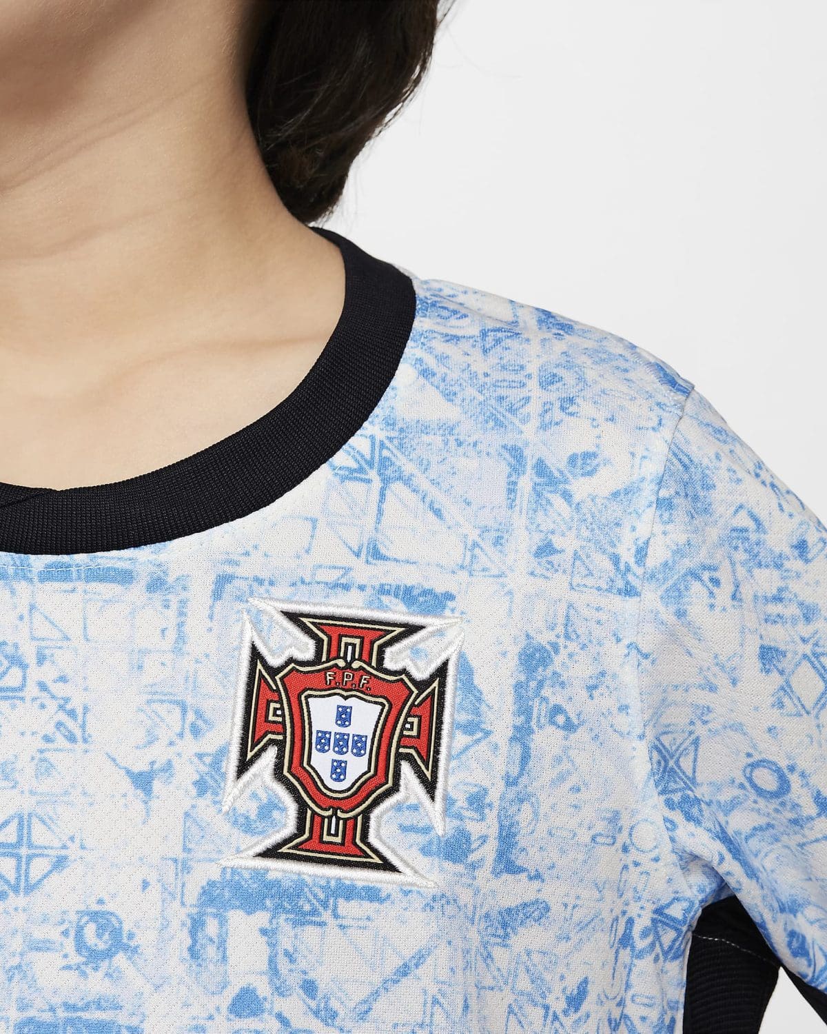 Close-up of a light blue jersey with a black collar and the Portugal crest displayed on the chest, featuring a geometric blue pattern in the background.