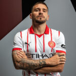 Portrait of a male football player with arms crossed, wearing a white jersey with red stripes and Girona FC crest.