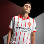 Young man wearing a white soccer jersey with red vertical stripes and Girona FC crest, posing in a studio.