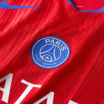 Close-up of a Paris Saint-Germain football jersey badge on red fabric with a blue Nike swoosh nearby.