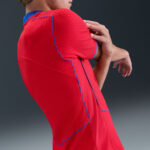 Person in a red athletic shirt with blue piping stretching shoulder backward, partial side view.