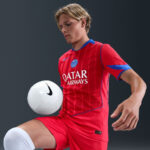 Soccer player in a red PSG jersey with Qatar Airways sponsor juggling a white ball against a dark background.