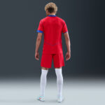 Back view of a soccer player in a red kit with blue trim, standing on a neutral gradient background