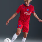 Soccer player in a red Paris Saint‑Germain kit, about to kick a white ball on a gray backdrop.