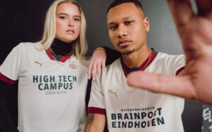 psv-away-23-24-puma-nss-14_84c1d0d8-9132-4a13-bf1e-dd26e8c336d5 | UAE SPORT STORE Two young people pose together in PSV Eindhoven jerseys; the woman rests her hand on the man's shoulder against a dark background.