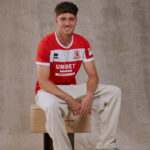 Portrait of a young man in a red and white soccer jersey seated on a wooden crate, smiling at the camera against a neutral backdrop.