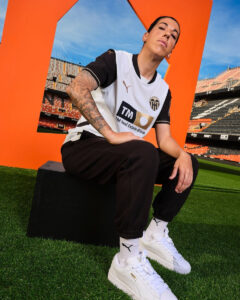 Athlete in Valencia CF jersey seated on a black block in a stadium with an orange architectural frame behind.