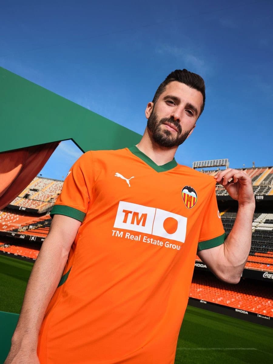 qBw3cxmb9QrnXQH | UAE SPORT STORE Man in an orange soccer jersey stands in a stadium, looking at the camera, with TM Real Estate sponsor on chest and Valencia crest visible.