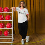 Person with curly hair wearing a white Liverpool FC jersey leans on a metal ball rack filled with red footballs in a gym with a yellow curtain behind him/her.