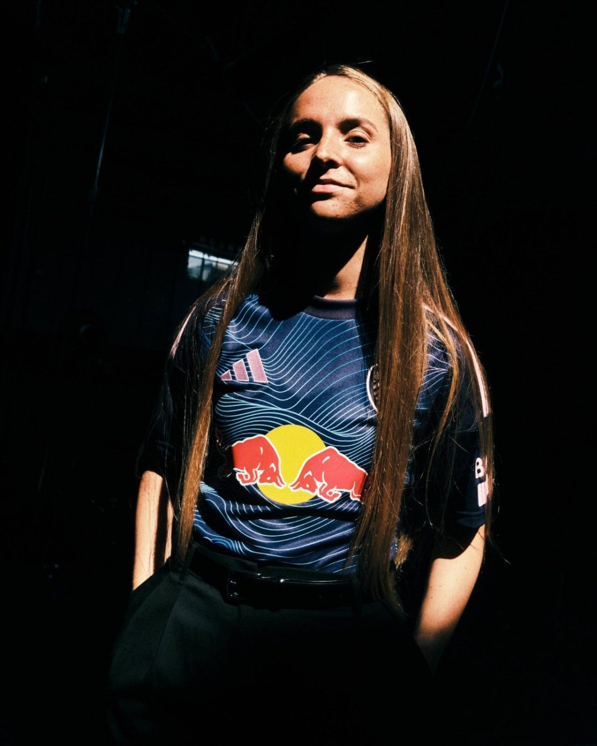 Portrait of a female athlete with long straight hair, wearing a blue Red Bull jersey under a spotlight in a dark setting.